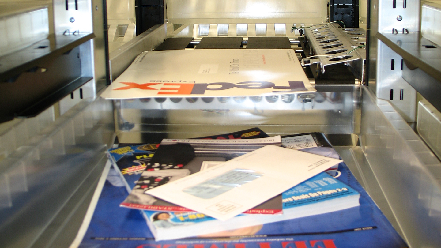 Stack of various mail items, including magazines, envelopes, and a FedEx package, inside a metal sorting machine.